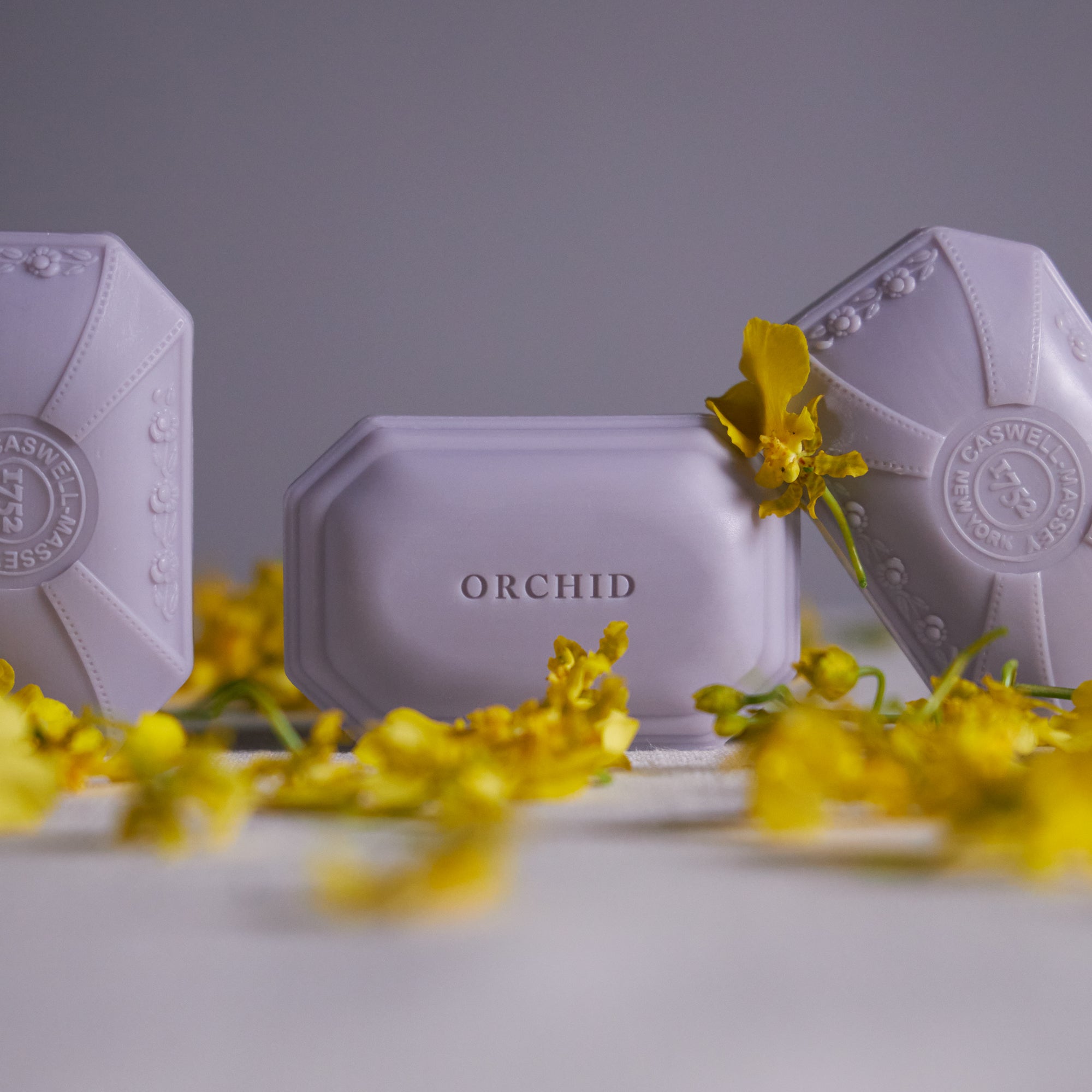 Caswell-Massey Orchid Bar Soap: Three bars of Orchid Soap in soft lavender color shown with orchid petals and flowers
