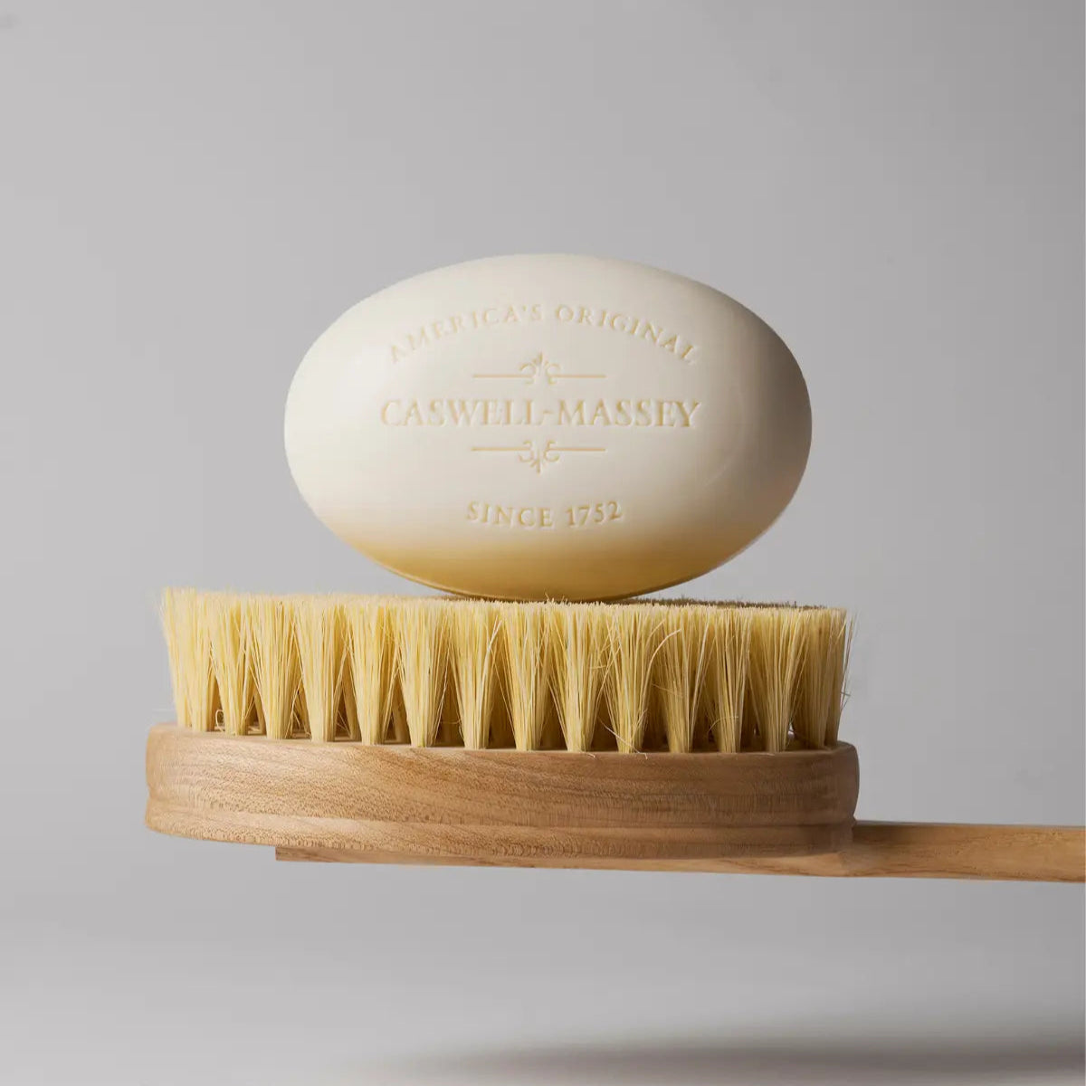 Caswell-Massey Almond Bar Soap white 5.8oz oval bar soap shown sitting atop a shower brush with wooden handle