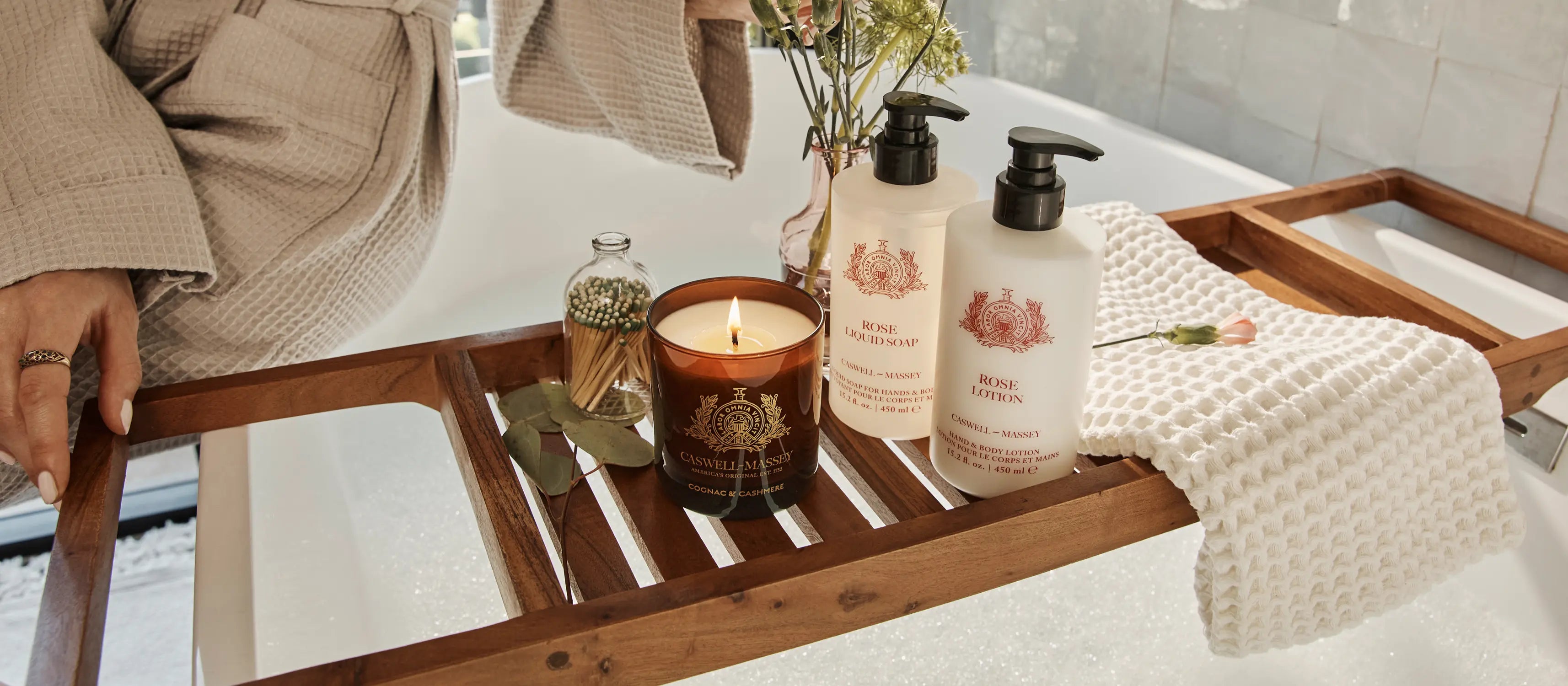 Caswell-Massey: Cognac & Cashmere Candle shown with Rose Lotion and Rose Liquid Soap on bath caddy sitting on drawn bathe as woman in robe readies to get in.