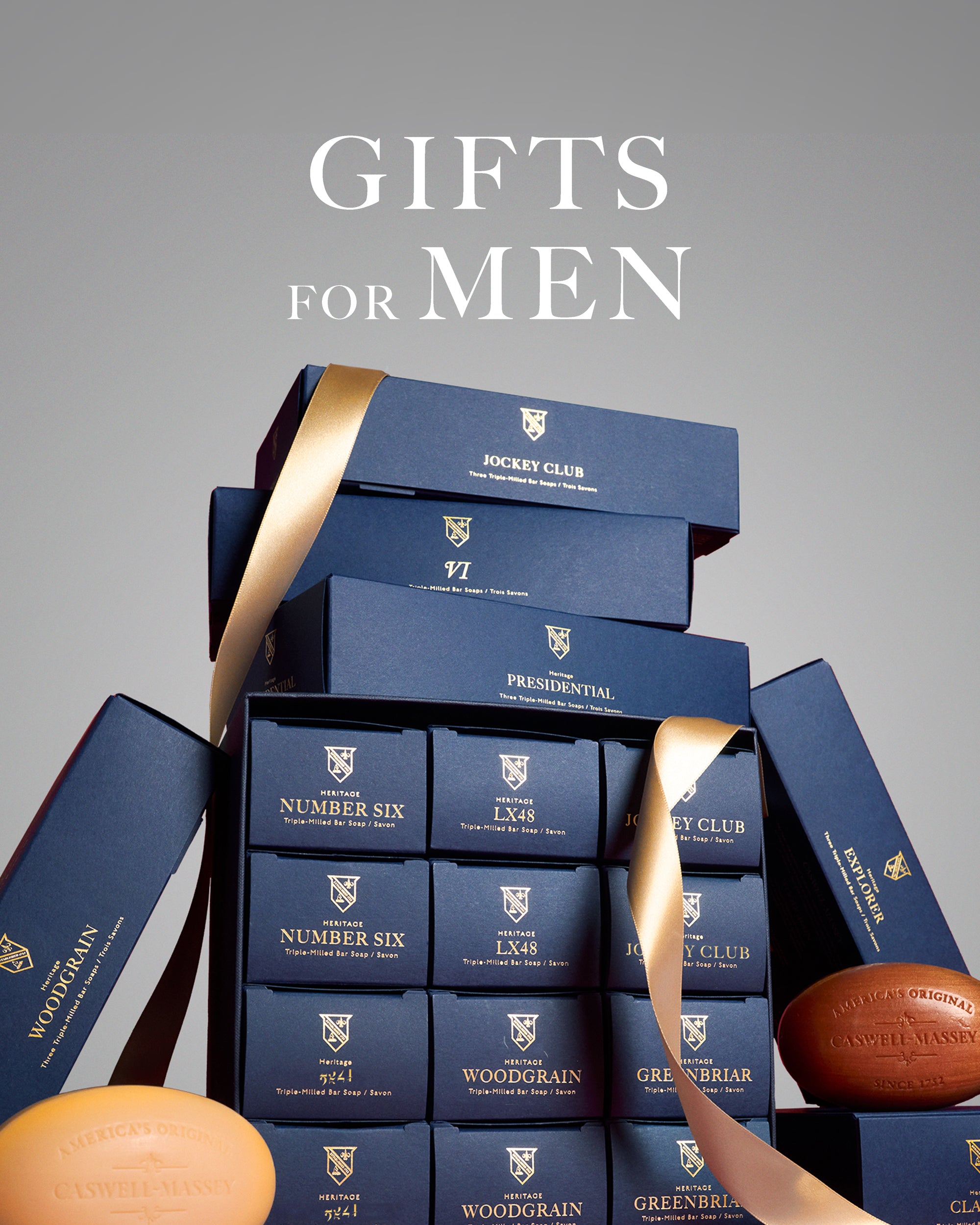 Caswell-Massey Gifts for Men. Image shows a stack of navy blue Caswell-Massey gift boxes that include gifts for men. Gifts include Presidential Soap Set, Jockey Club 3-Soap Set, and Number 3-Soap Set. Woodgrain bar soap and Number Six bar soap, both unwrapped, can be seen leaning against Heritage Year of Soap gift set.