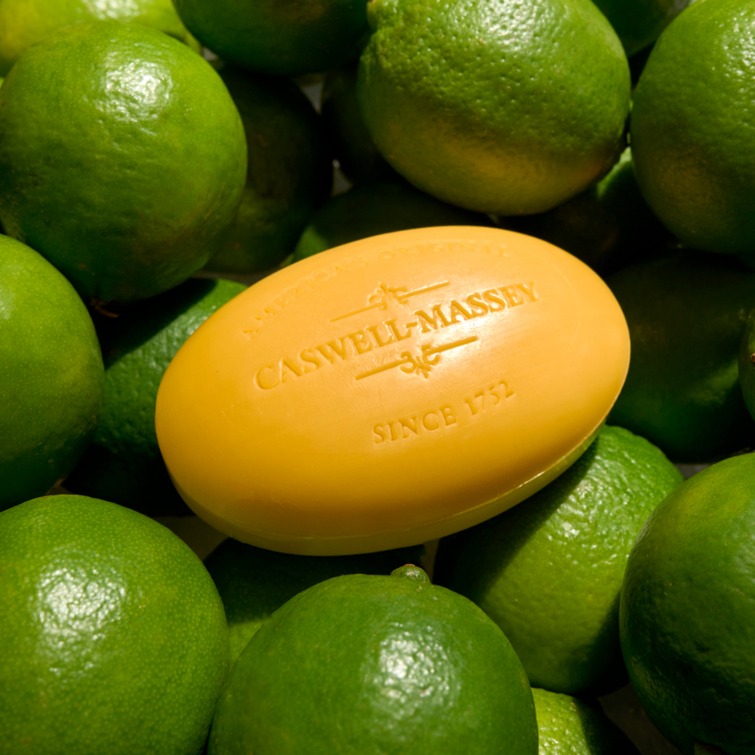A round yellow bar of Caswell-Massey's Verbena Bar Soap embossed with the brand name 'CASWELL-MASSEY' and 'SINCE 1732', resting on a bed of fresh green limes.