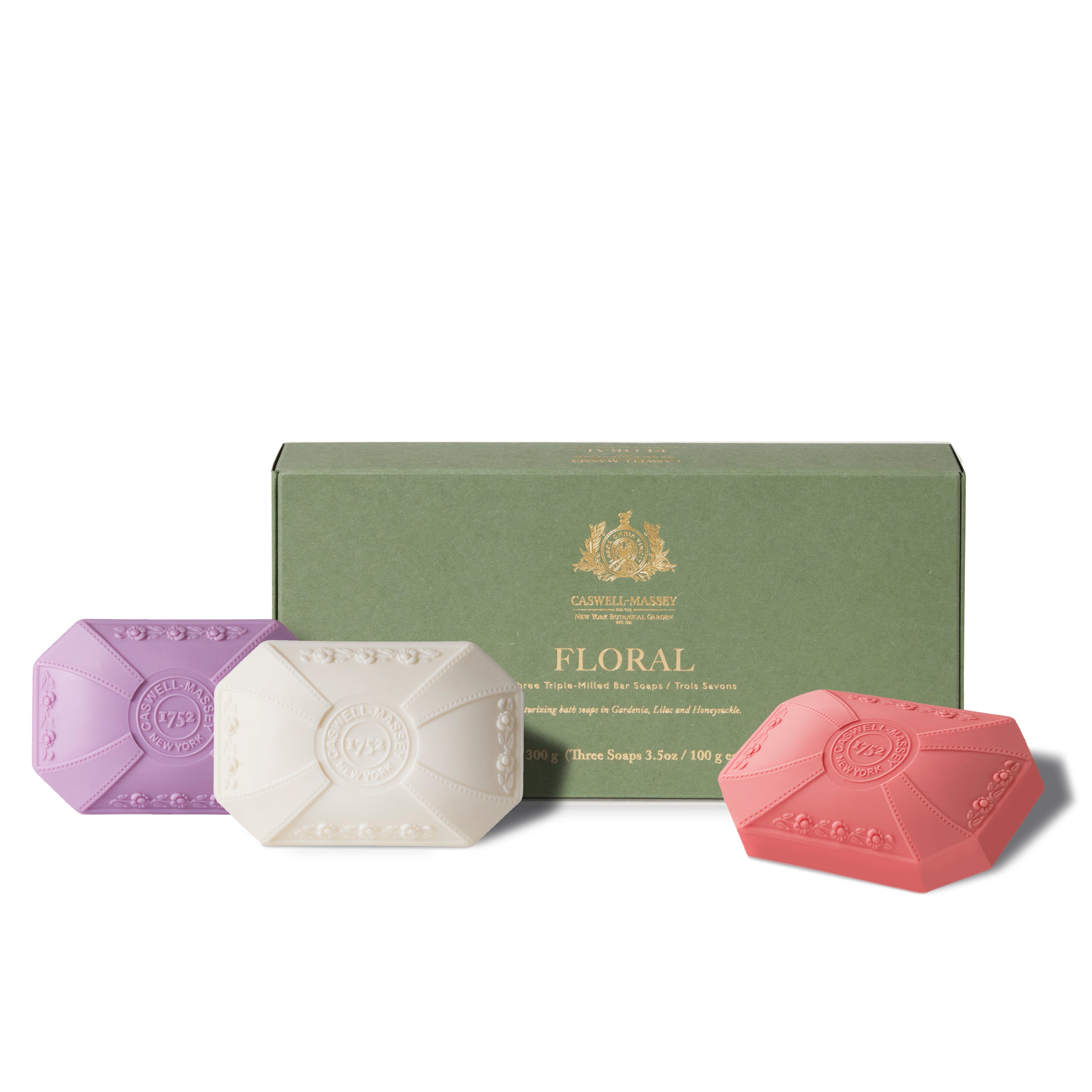 Caswell-Massey® Trio of Living Florals Three-Soap Set: three bath soaps shown sitting in front of soft green gift box (packaging). One purple soap for Lilac, one white for Gardenia, and one mauve for Honeysuckle soap. All featuring a decorative molded shape. 