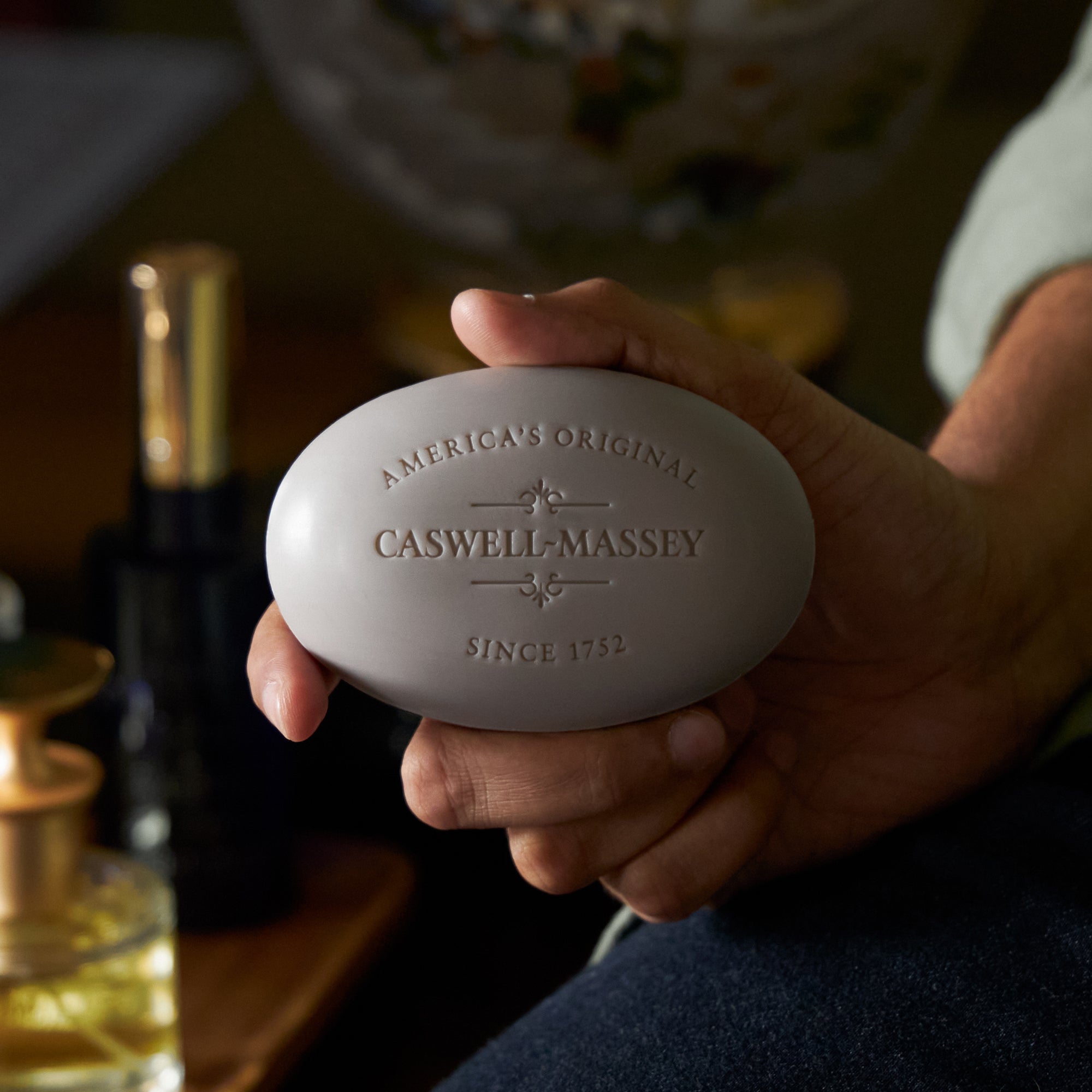 Caswell-Massey LX48 Bar Soap: Man holding the oval bar soap next to fragrance bottles