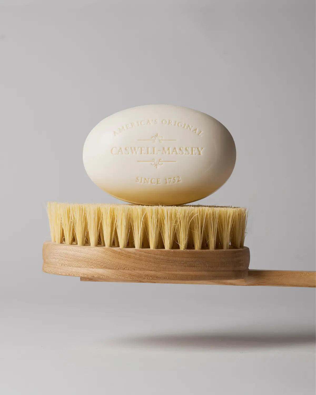 Caswell-Massey Almond Bar Soap white 5.8oz oval bar soap shown sitting atop a shower brush with wooden handle