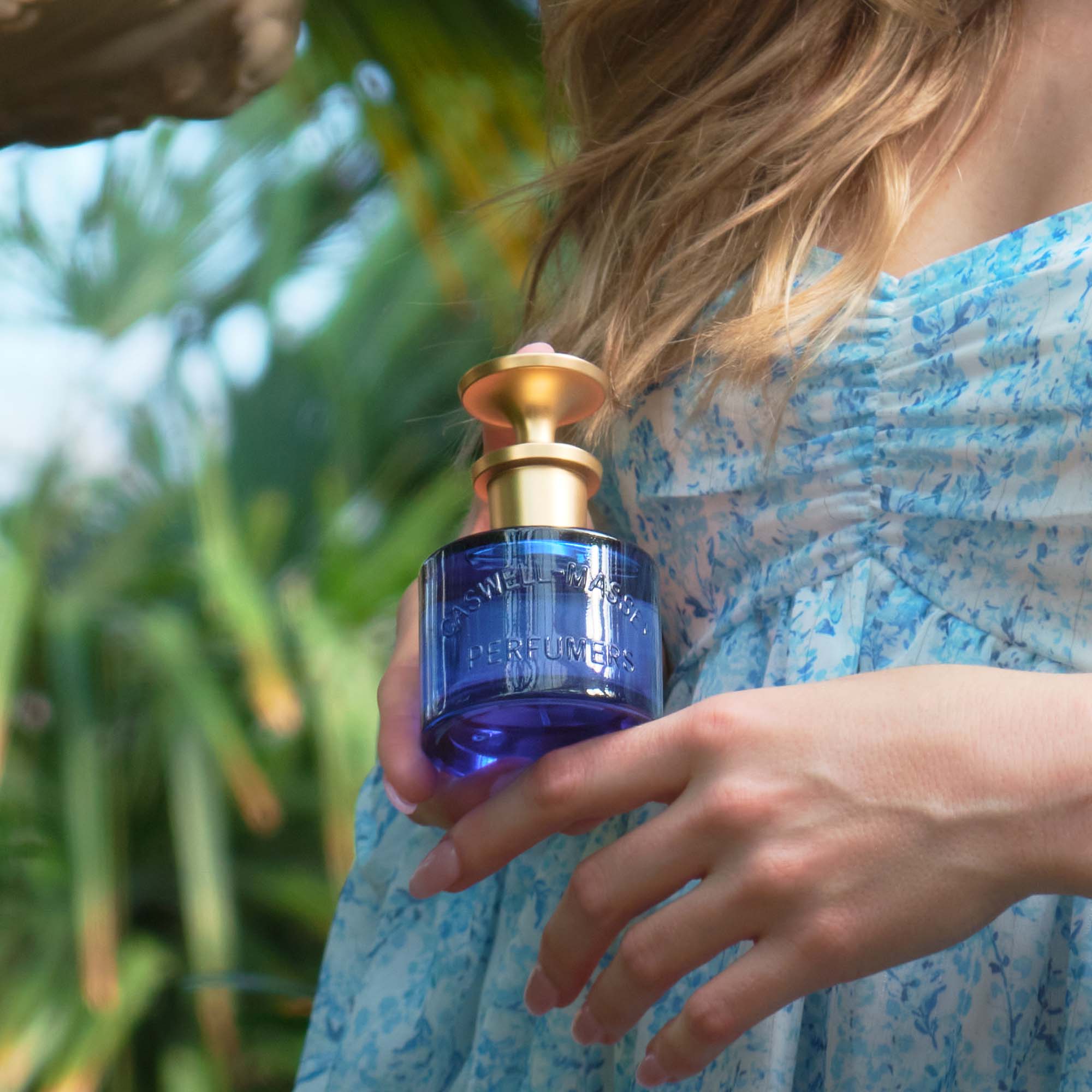 Caswell-Massey Elixir of Love Eau de Toilette (60 mL), woman holding the fragrance bottle, cobalt blue with gold cap, in her hands in front of a floral sundress with soft blue floral pattern