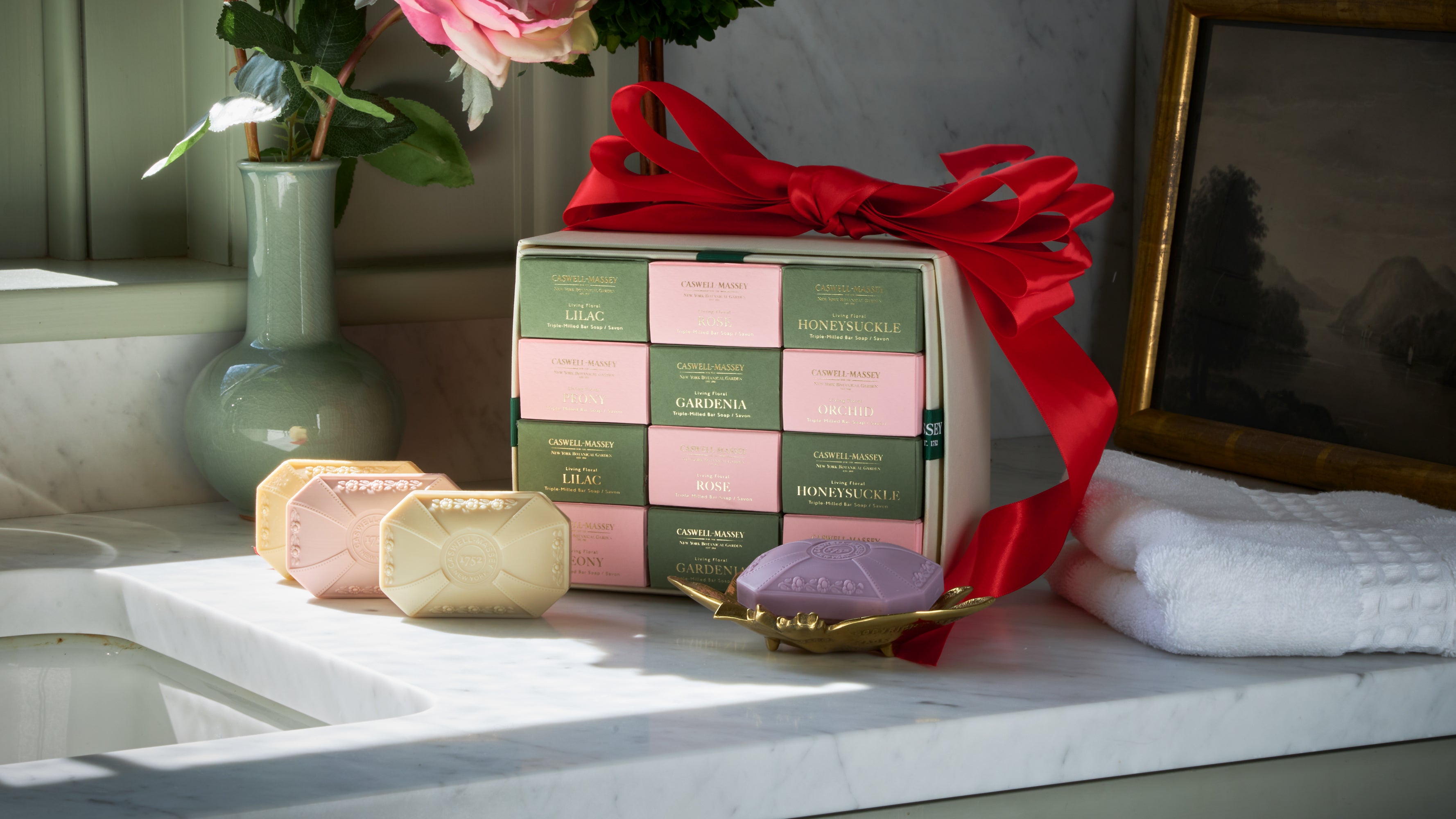 Caswell-Massey's Designer Floral Twelve Bar Soap gift set sitting on a white granite counter top with red ribbon draped across the box and soaps laying on the counter in front of the box 