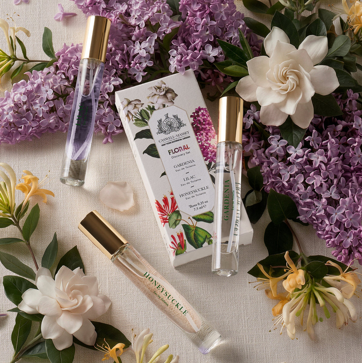 Caswell-Massey Floral Discovery Set: featuring Lilac Eau de Toilette, Gardenia Eau de Toilette, and Honeysuckle Eau de Toilette. Shown surrounded by the namesake flowers.