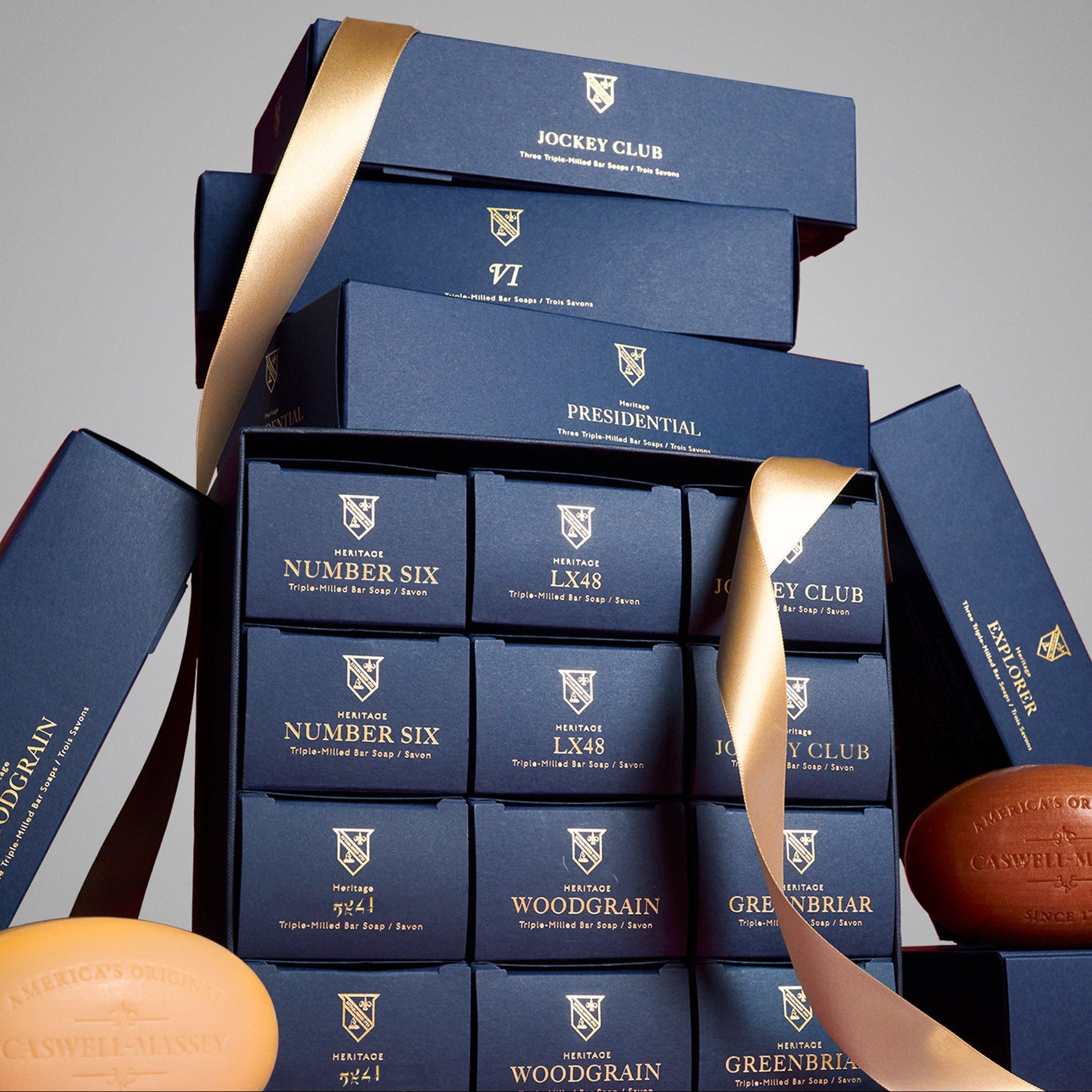 Caswell-Massey Gifts for Men. Image shows a stack of navy blue Caswell-Massey gift boxes that include gifts for men. Gifts include Presidential Soap Set, Jockey Club 3-Soap Set, and Number 3-Soap Set. Woodgrain bar soap and Number Six bar soap, both unwrapped, can be seen leaning against Heritage Year of Soap gift set.