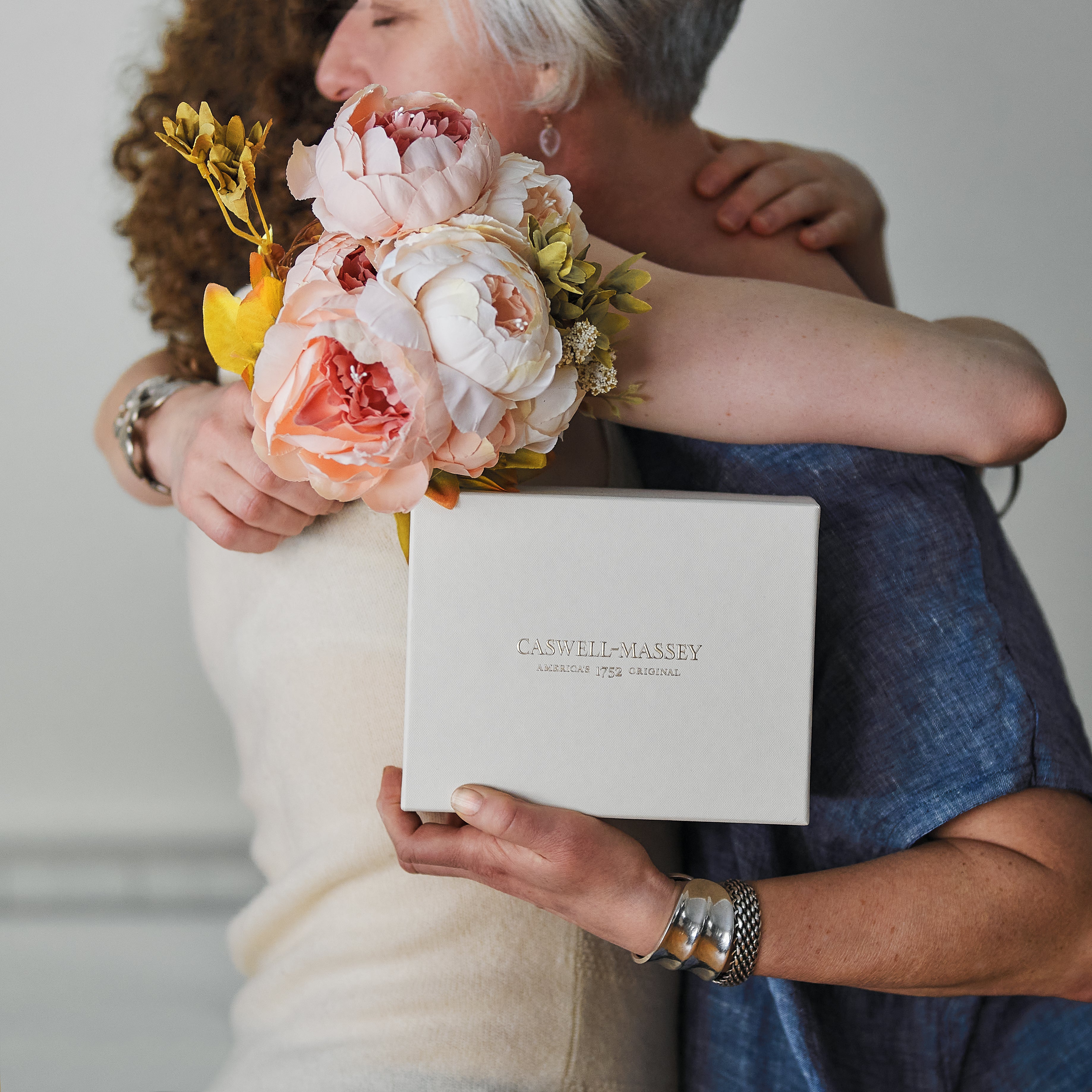 Mother hugging daughter as mother accepts Caswell-Massey Gift Set and bouquet of flowers