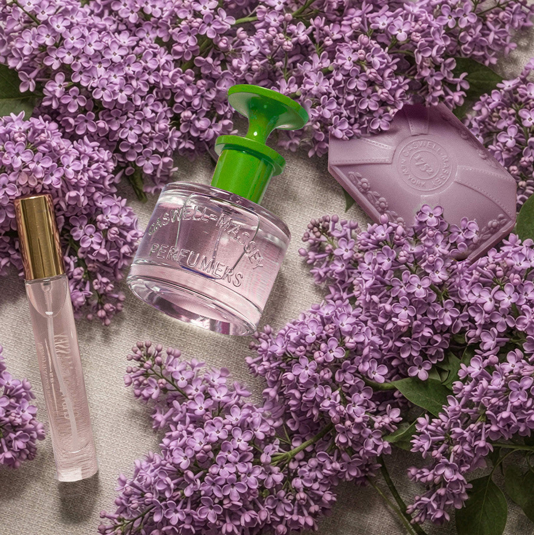 A Lilac Eau de Toilette perfume bottle with a bright green cap and a clear glass body, surrounded by purple lilac flowers on a light beige fabric background. A lavender soap bar and a clear glass roller bottle with a gold cap are also arranged around it.